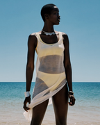 Woman in a sheer white dress standing on a beach with clear blue sky and water.