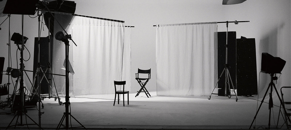 A black and white studio setup with white backdrop curtains, lighting stands, softboxes, and two empty chairs in the center of the set.