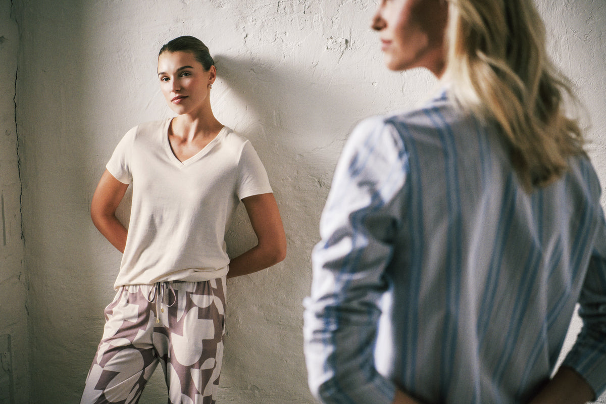 A woman stands against a textured white wall wearing a white V-neck short-sleeve top and patterned lounge pants. Another woman in a blue striped shirt is blurred in the foreground, creating a mirror-like effect.