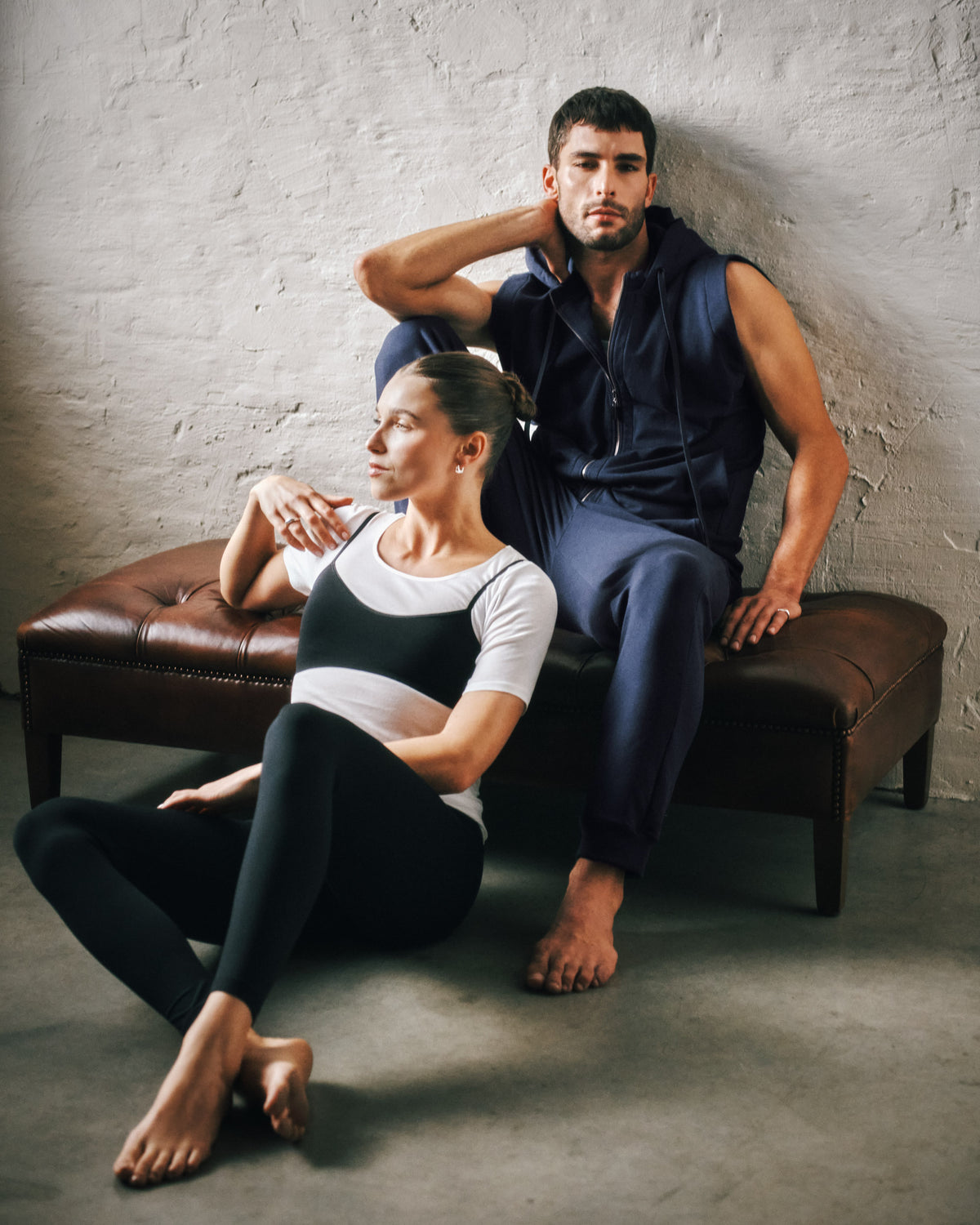 A man and a woman pose in matching black and white activewear sets in a studio setting with a textured gray wall and a brown leather bench.