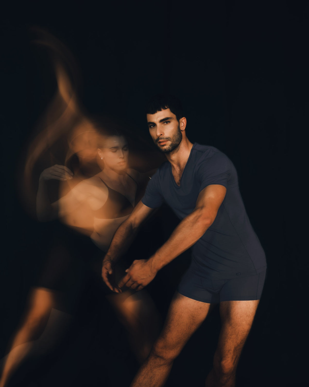 A man in a dark navy blue v-neck t-shirt and matching shorts poses dynamically against a dark background, with a motion-blurred duplicate figure in the background suggesting movement or dance.