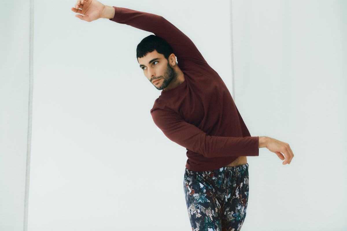 A man in a studio setting, wearing a long-sleeve maroon shirt and dark patterned pants, performing a side stretch with one arm extended overhead against a plain white background.