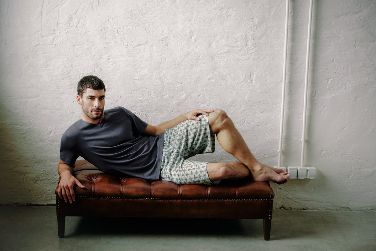 A man reclining on a brown leather bench against a textured white wall, wearing a dark gray t-shirt and patterned shorts.
