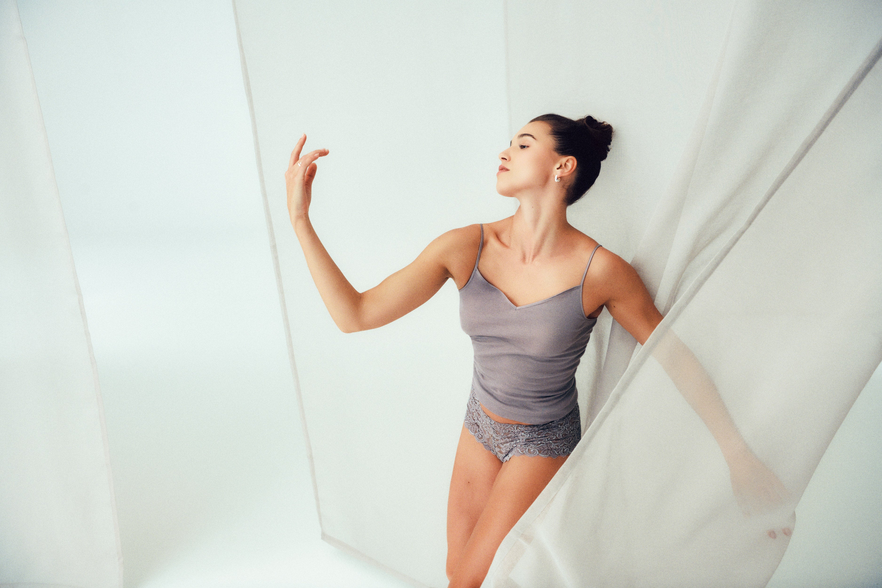 A woman modeling a gray camisole and matching lace panties while leaning against a white draped fabric backdrop in a studio setting.