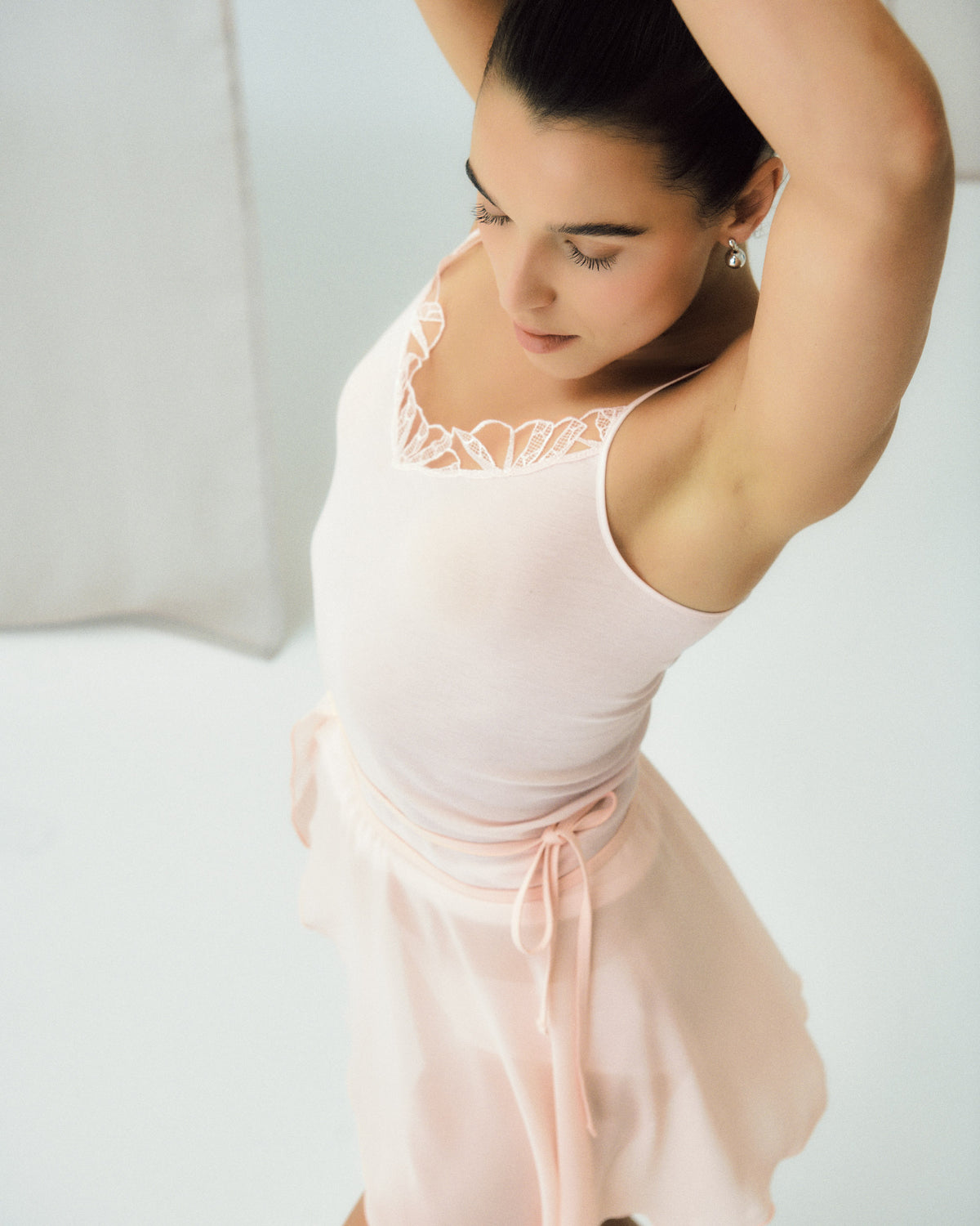 A woman wearing a light pink sleeveless camisole top with lace trim and a matching sheer pink wrap skirt, photographed in a bright studio setting.