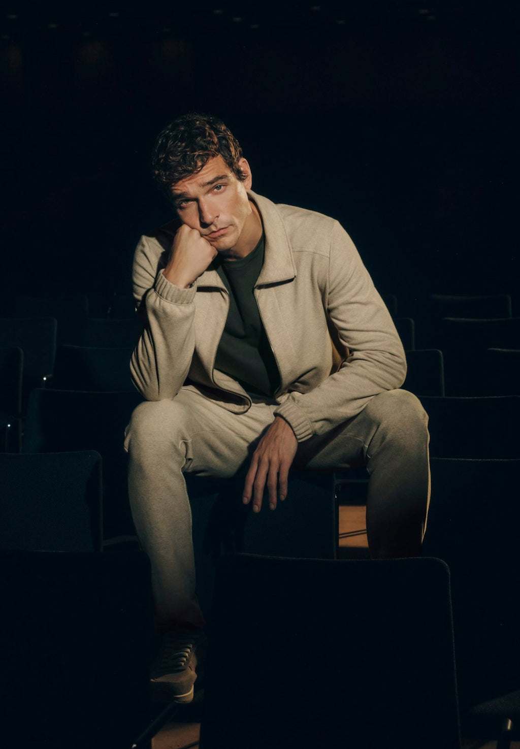 Man sitting in a dark theater setting wearing a beige outfit.