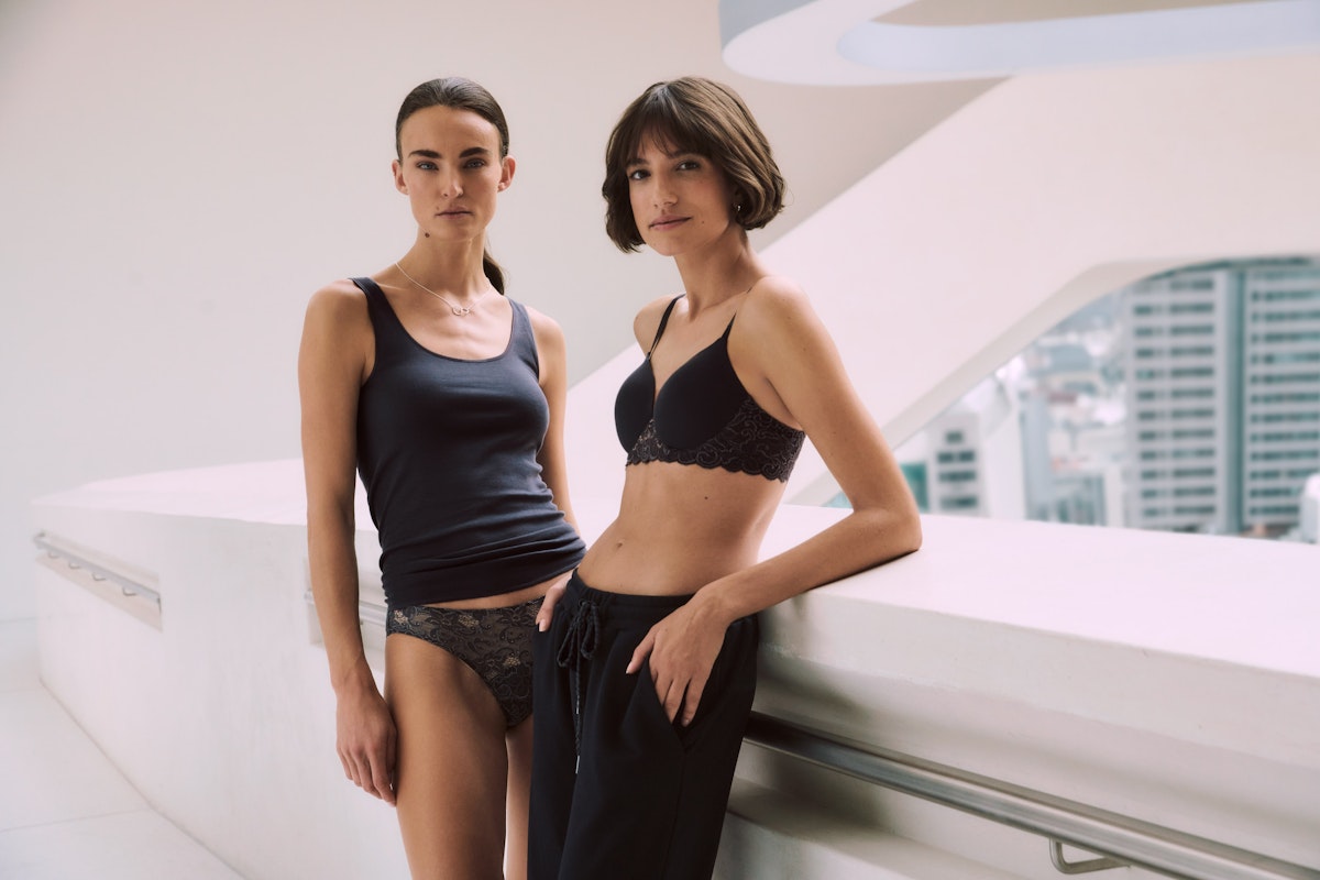 Two women in black lingerie standing on a white surface with a cityscape background
