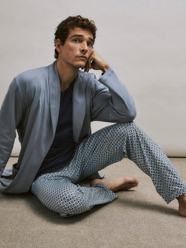 Man wearing a light blue robe and patterned pants sitting on the floor against a plain wall.