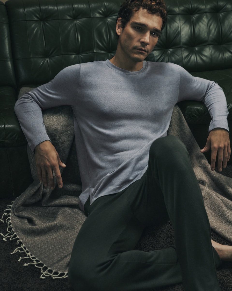 Man sitting on a dark leather couch wearing a light gray sweater and dark pants.