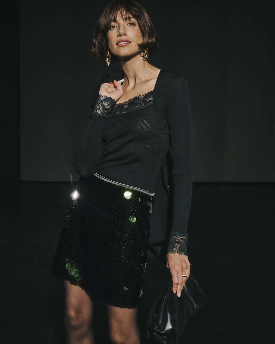 Woman wearing a dark green top with lace details and a black sequin skirt against a dark background.