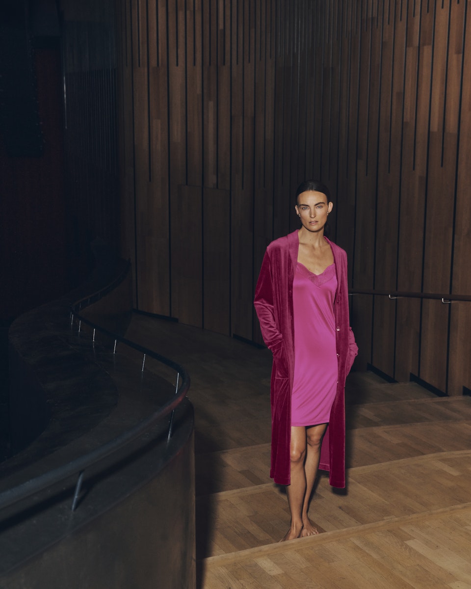 Woman wearing Lilith pink dress and Favourites robe standing in a dimly lit room with wooden walls.