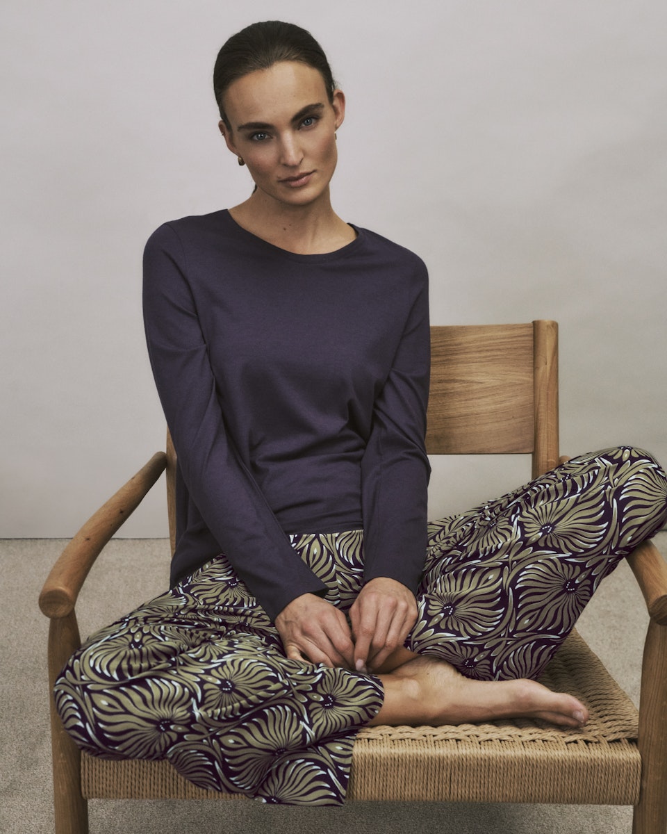 Woman sitting on a wooden chair wearing a dark long-sleeve top and patterned pants.