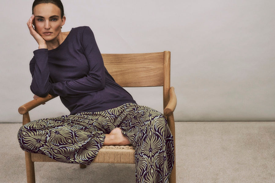Woman sitting on a wooden chair wearing a dark blue top and patterned pants.