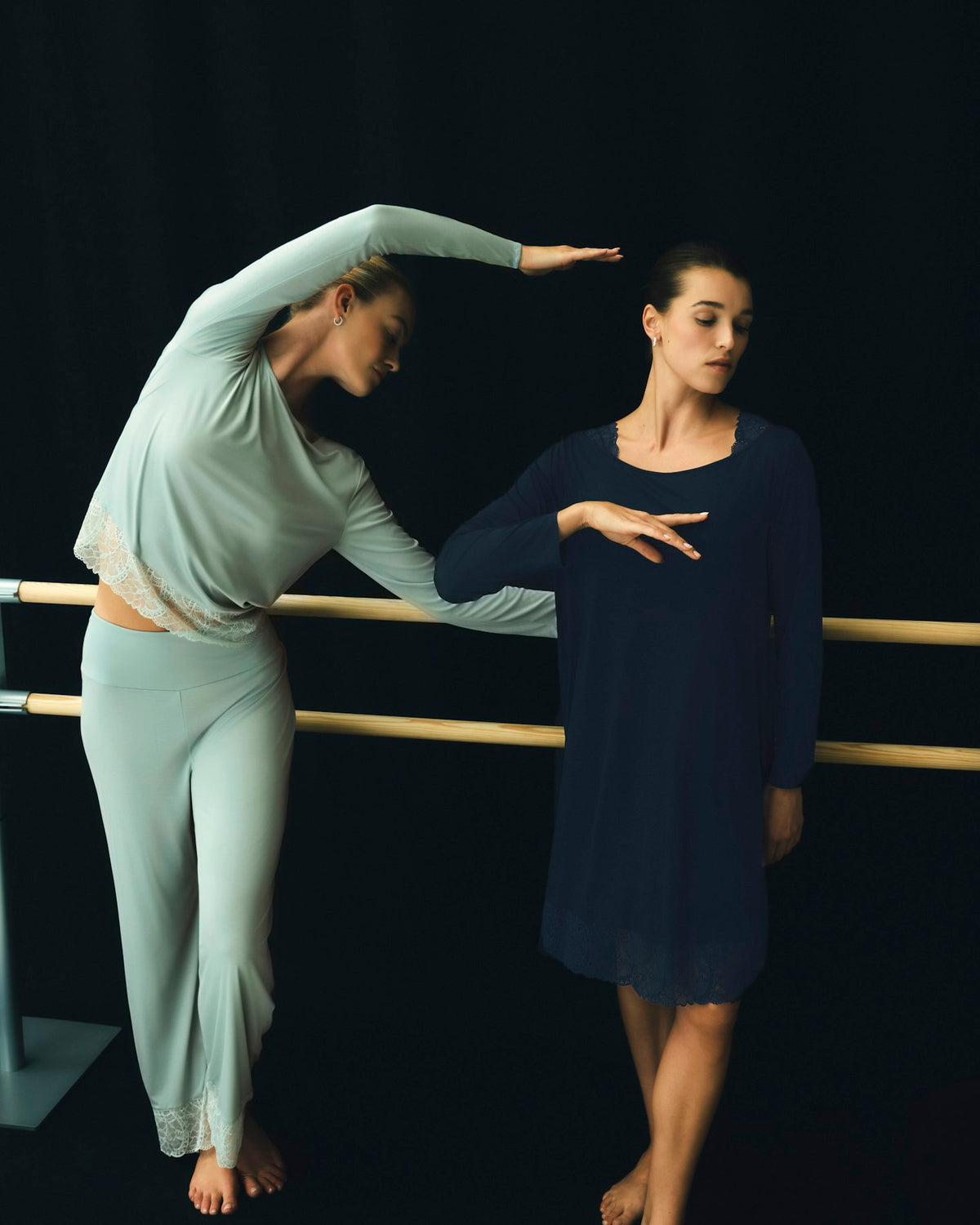 Two women in dancewear pose in front of a black studio background with a wooden barre. One wears a pale mint green long-sleeve crop top and matching pants, while the other wears a dark navy blue long-sleeve dress.