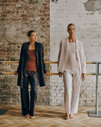 Two models standing in a studio with a brick and white plaster wall background, wearing matching lounge sets. The model on the left wears a dark navy patterned blazer and trousers over a rust-brown top.