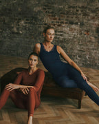 Two women modeling matching athletic sets in a rustic indoor setting with a brick wall and wood floor. One woman sits on a dark leather bench wearing a rust-brown long-sleeve top and leggings set, while the other reclines on the bench wearing a navy blue lace-trim tank top and leggings set.