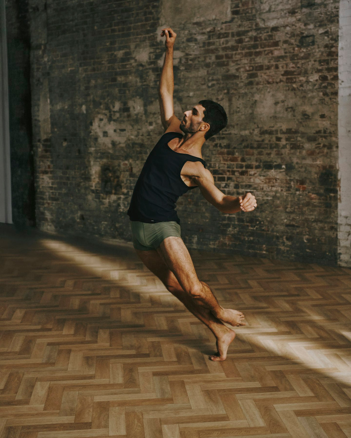 A man in a black tank top and olive green shorts poses dynamically in a dance or yoga pose within a rustic room featuring a brick wall and a herringbone wood floor.