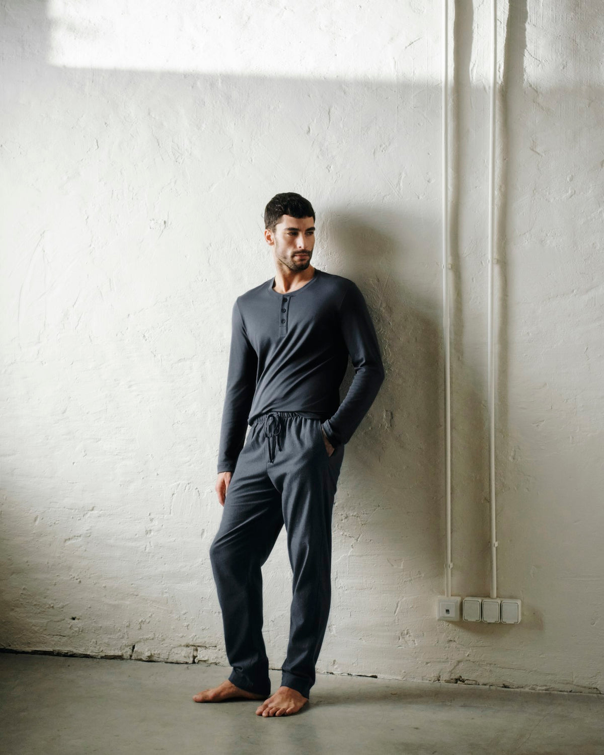 A man wearing a dark gray long-sleeve henley shirt and matching dark gray sweatpants stands barefoot against a textured white plaster wall.