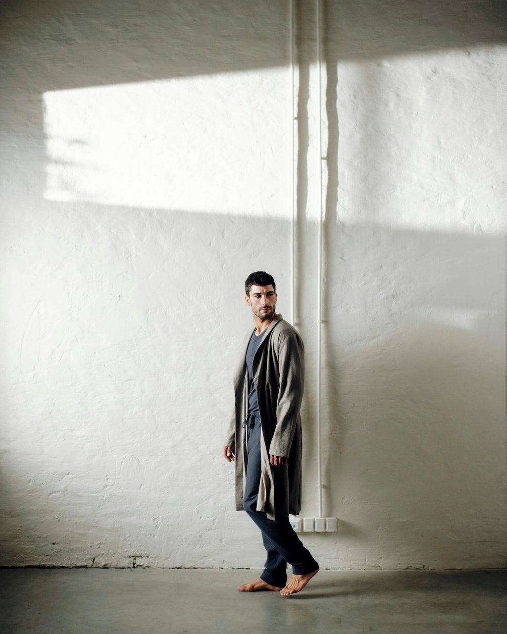 A man stands barefoot against a textured white wall in a minimalist room, wearing a long gray robe over a shirt and dark trousers.