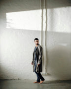 A man stands barefoot against a textured white wall in a minimalist room, wearing a long gray robe over a shirt and dark trousers.