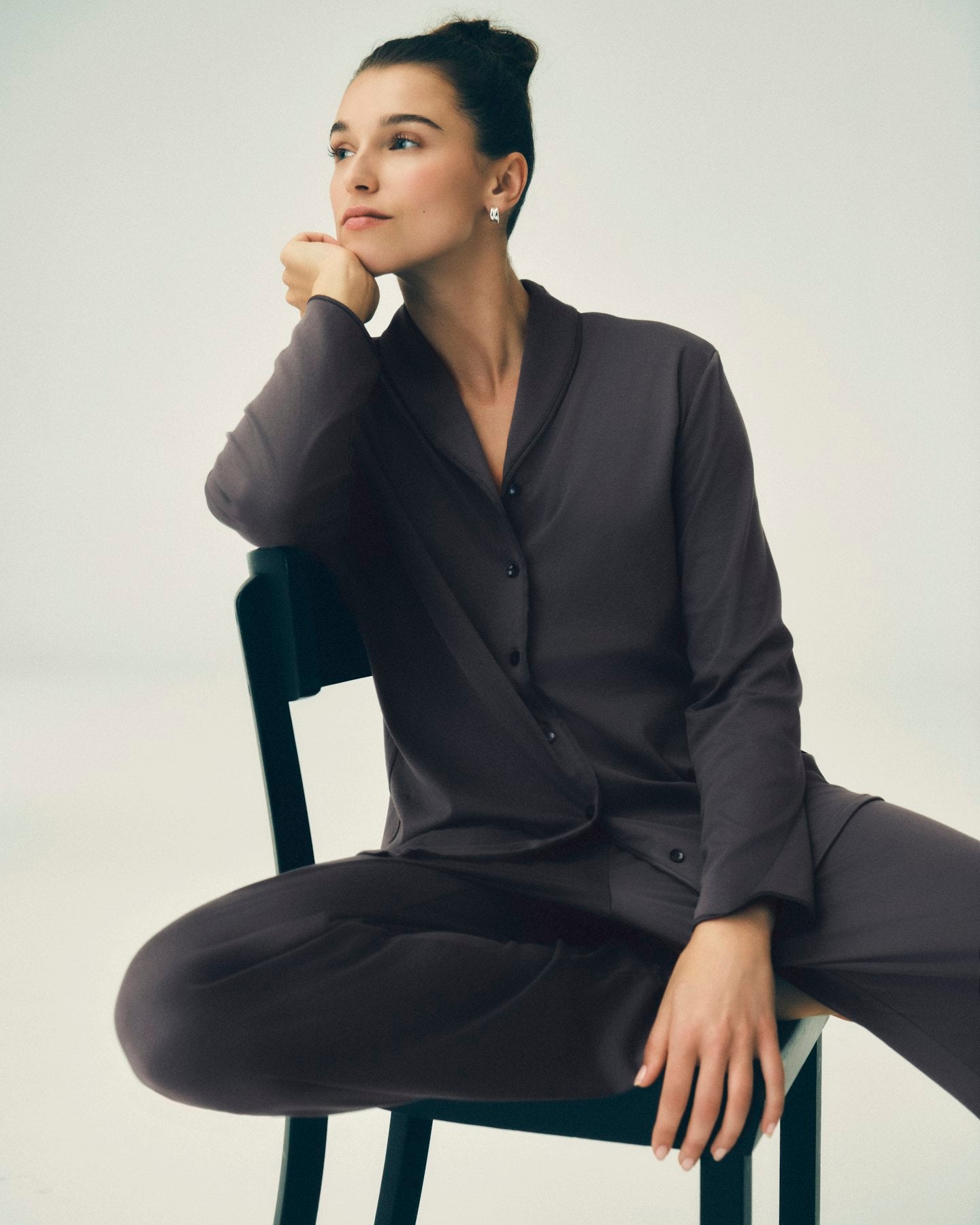 A woman wearing a dark charcoal gray pajama set with a button-front long-sleeve top and matching pants, seated on a black chair against a light gray studio background.