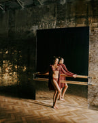 A female dancer in matching burgundy and brown dancewear pose in front of a large black mirror in a rustic studio with brick walls and wooden floors.