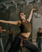 A woman in a dance studio wearing a brown ribbed tank top and dark pants, captured mid-motion with arms extended. Other dancers are blurred in the background against a textured concrete wall.