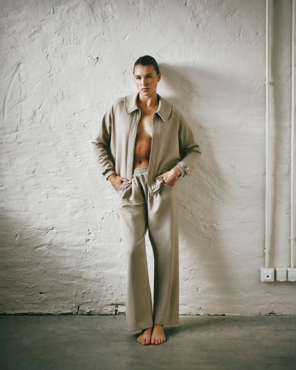 A model stands barefoot against a textured white plaster wall, wearing a matching beige two-piece outfit consisting of zip-up jacket and wide-leg trousers.