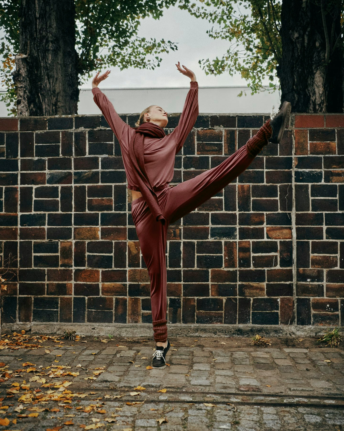 A person in a matching rust-brown tracksuit poses dynamically with one leg extended upward in front of a dark brick wall outdoors.