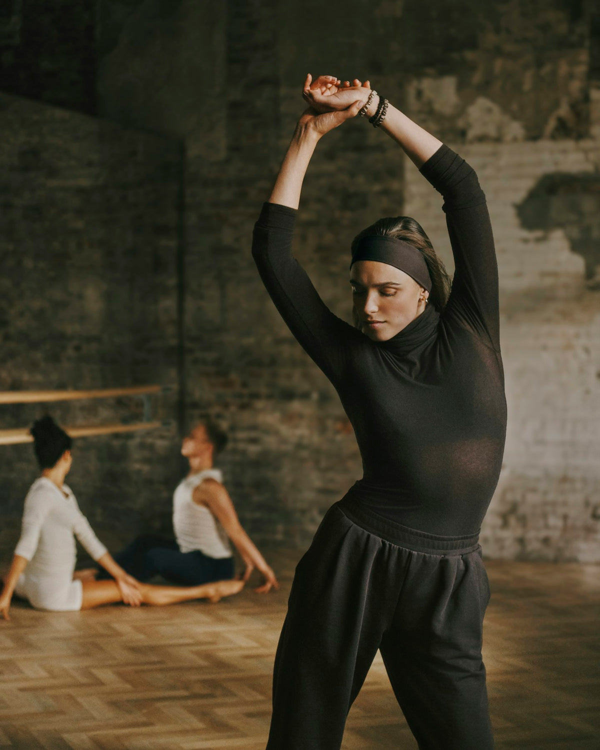 A woman in a black long-sleeve mock neck top and black sweatpants stretches her arms overhead in a rustic studio with brick walls and wood floors. Two other dancers are seated in the background.