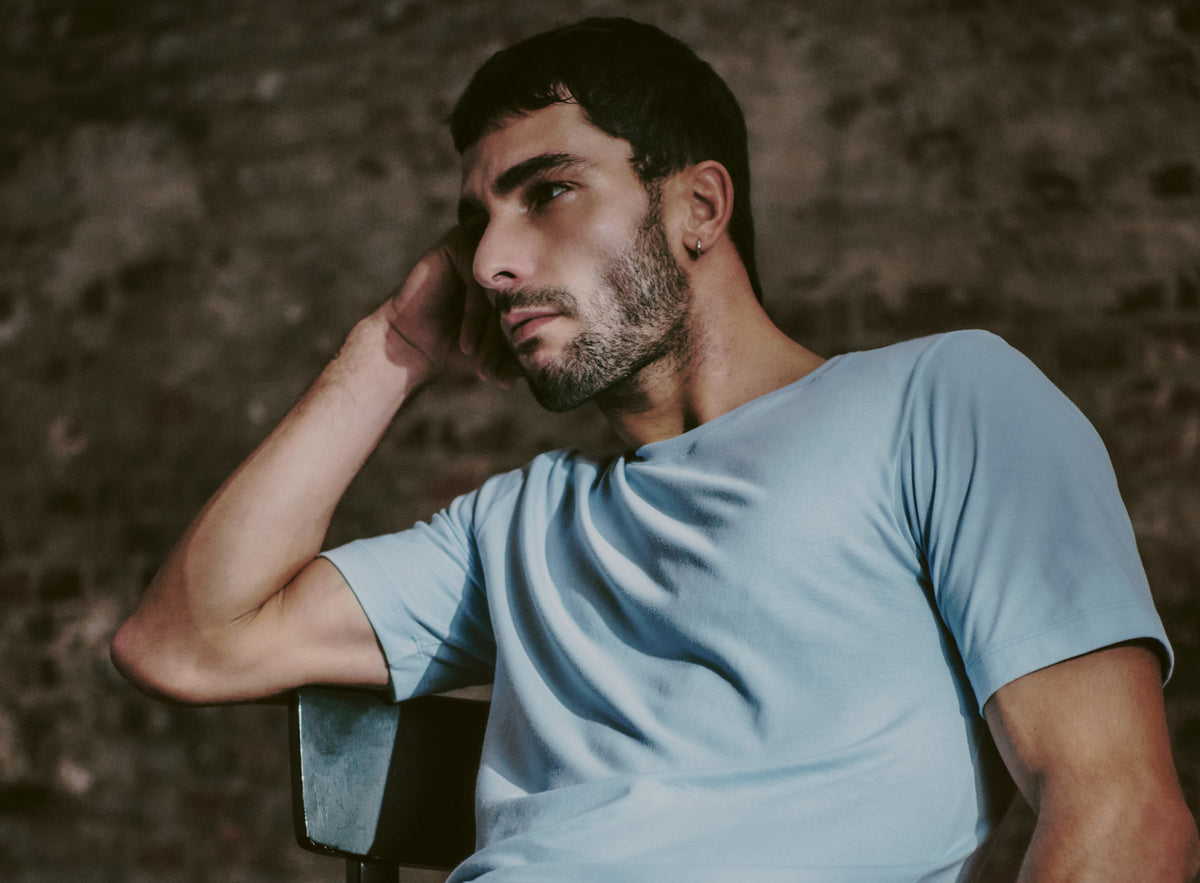 A man wearing a light blue crew-neck t-shirt, sitting against a dark textured brick wall background.