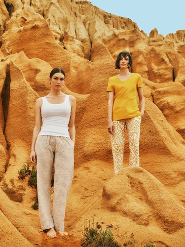 Two women standing on sand dunes. Women in front is wearing Sleep And Lounge Ribbed Cotton Tank Top in White with Sleep And Lounge Cotton Pajama Pants in Raffia Stripe. The women standing in back is wearing Sleep And Lounge Relaxed V-Neck T-Shirt in Sun Kiss with Sleep And Lounge Cotton Blend Drawstring Pants in Golden Ornament