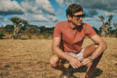 Man in sunglasses, looking in the grass, holding small flowers, wearing Living Shirts in Chutney with Night and Day Woven Shorts in Bluewood Strip.