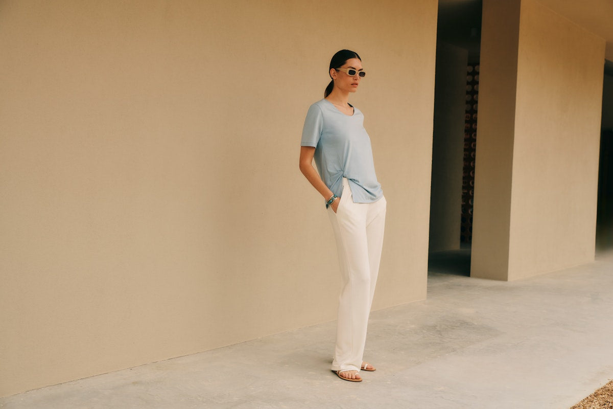 Woman standing outside, wearing the Natural Shirt in Sea Breeze with Pure Comfort Pants in Misty White, paired with sunglasses.