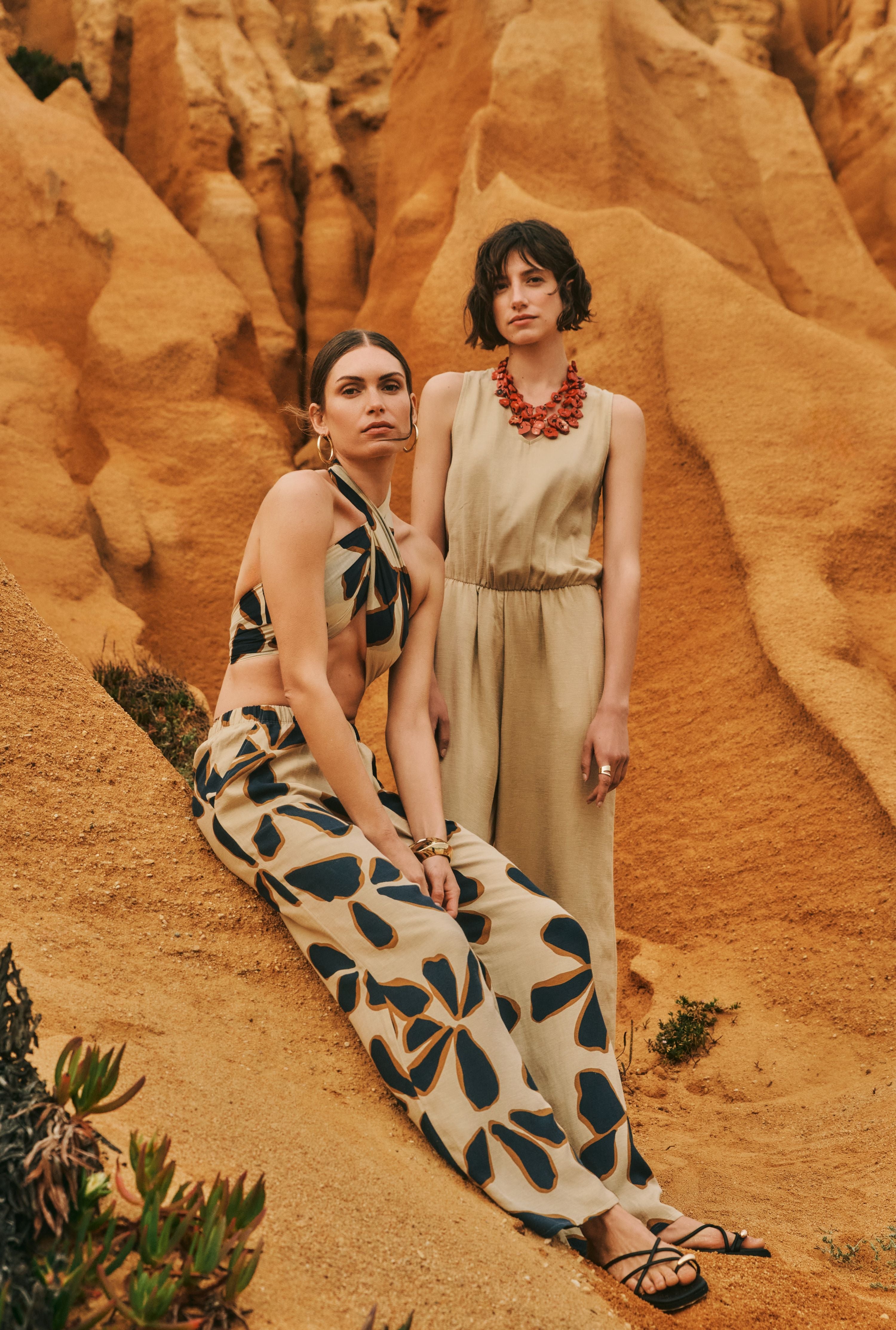 Two women together on sand dunes. Women sitting down on the left is wearing Accessories Cotton Pareo tied as a top, paired with Urban Casuals Linen Blend Drawstring Pants in Desert Flower. Women standing to the right is wearing Urban Casuals Linen Blend Jumpsuit in Iced Cappuccino