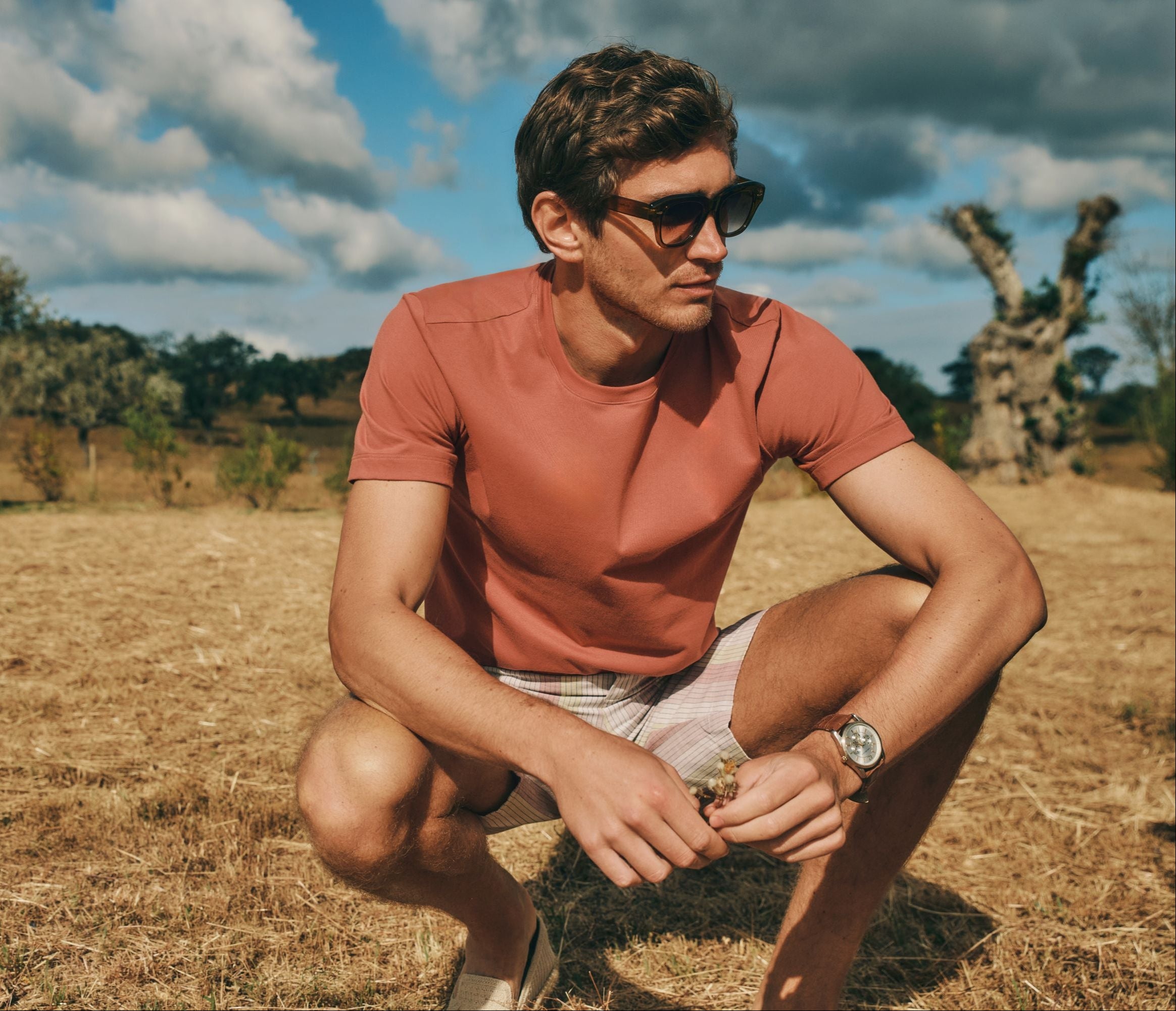 Man in sunglasses, looking in the grass, holding small flowers, wearing Living Shirts in Chutney with Night and Day Woven Shorts in Bluewood Stripe