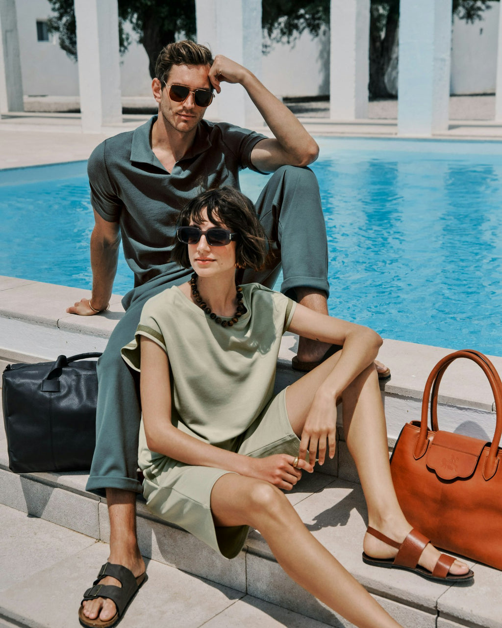 Man and woman sitting by a poolside with sunglasses and handbags.