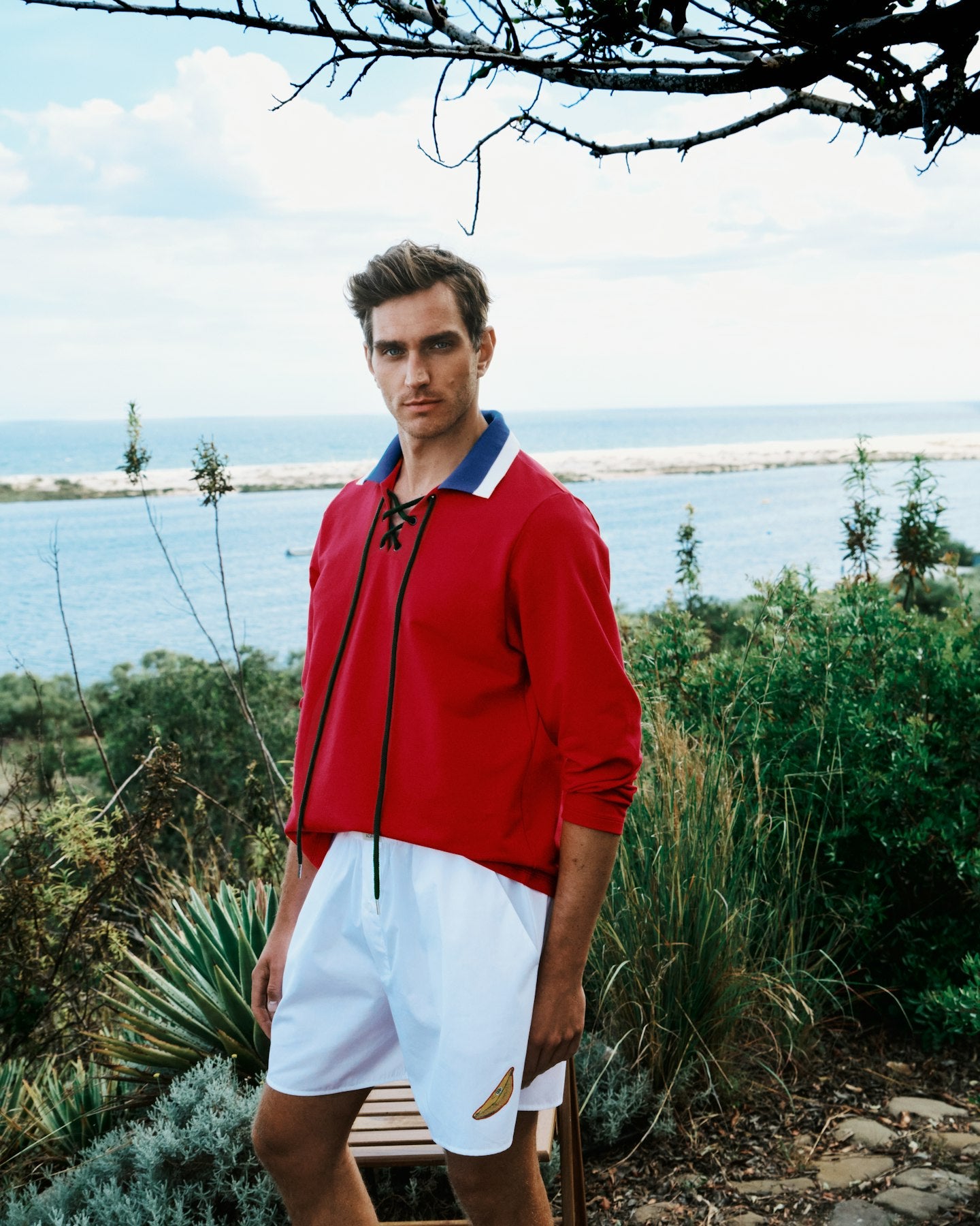 Man wearing a red shirt and white shorts standing outdoors with ocean and sky in the background