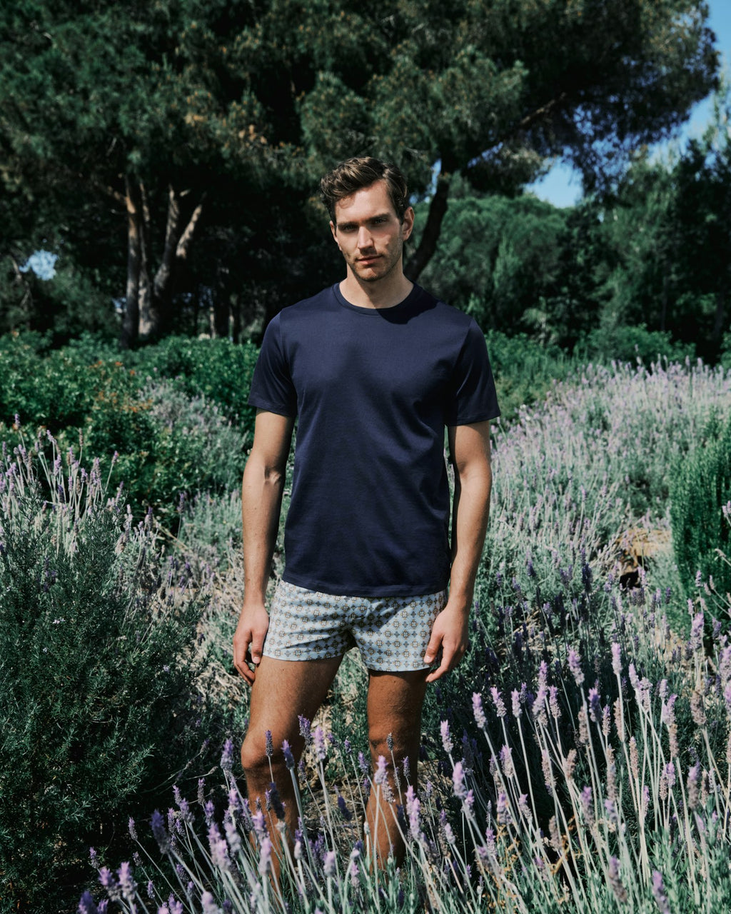 Man standing in a lavender field wearing a navy t-shirt and patterned shorts.