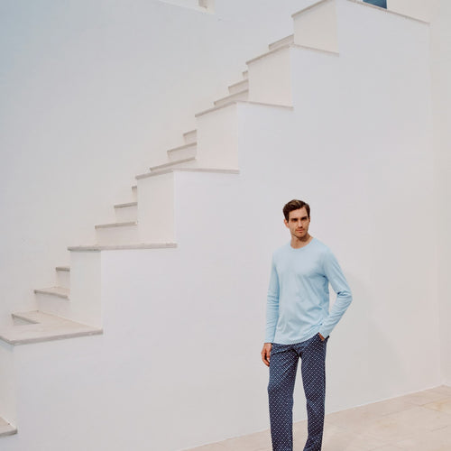 Man wearing a light blue long sleeved top and patterned pants standing in front of a white staircase.