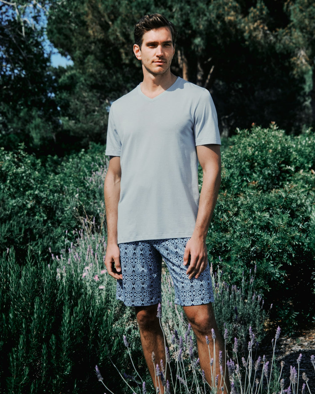 Man wearing a light gray t-shirt and patterned shorts standing in a garden with greenery and lavender.