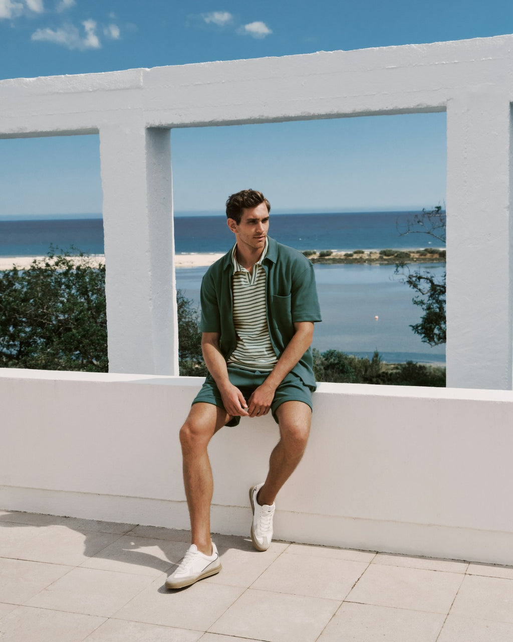 Man wearing a green outfit sitting on a white railing overlooking a scenic view of the ocean.