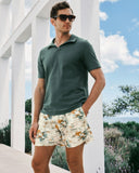 Man wearing a green polo shirt and floral shorts standing outdoors with white columns and blue sky in the background.