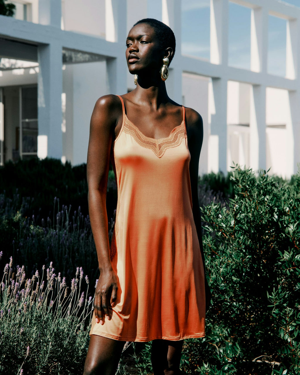 Woman wearing an orange dress standing outdoors with greenery and a building in the background