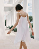 Woman in a white slip dress walking in a sunlit outdoor setting with columns and greenery.