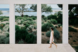 Woman walking on a path with a scenic view of trees and water through large windows.