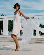 Woman in a light pink dress with a white scarf standing on a bridge with a blue sky background