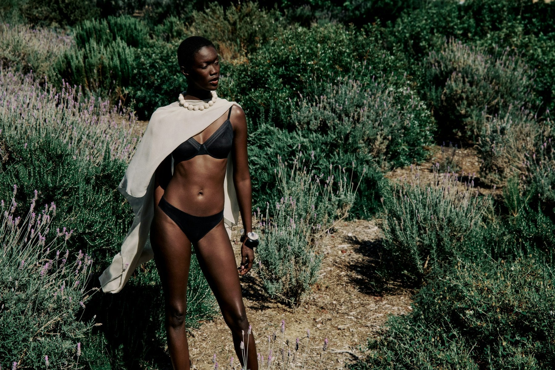 Woman in a black bikini and white shawl standing in a natural setting with greenery and lavender.