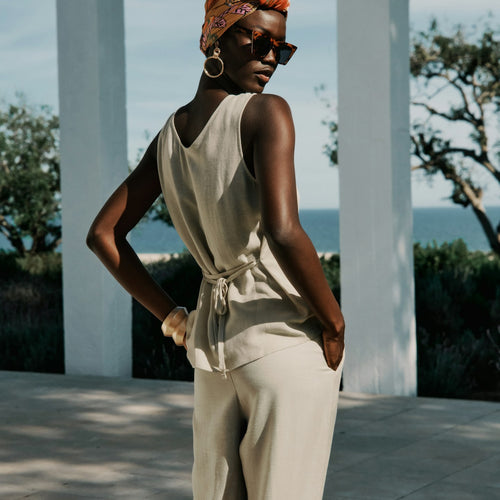 Woman in a beige outfit with a headscarf and sunglasses standing outdoors with ocean view.