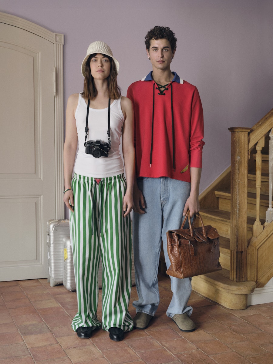 Two people standing in a room with a staircase, one holding a camera and the other a bag.